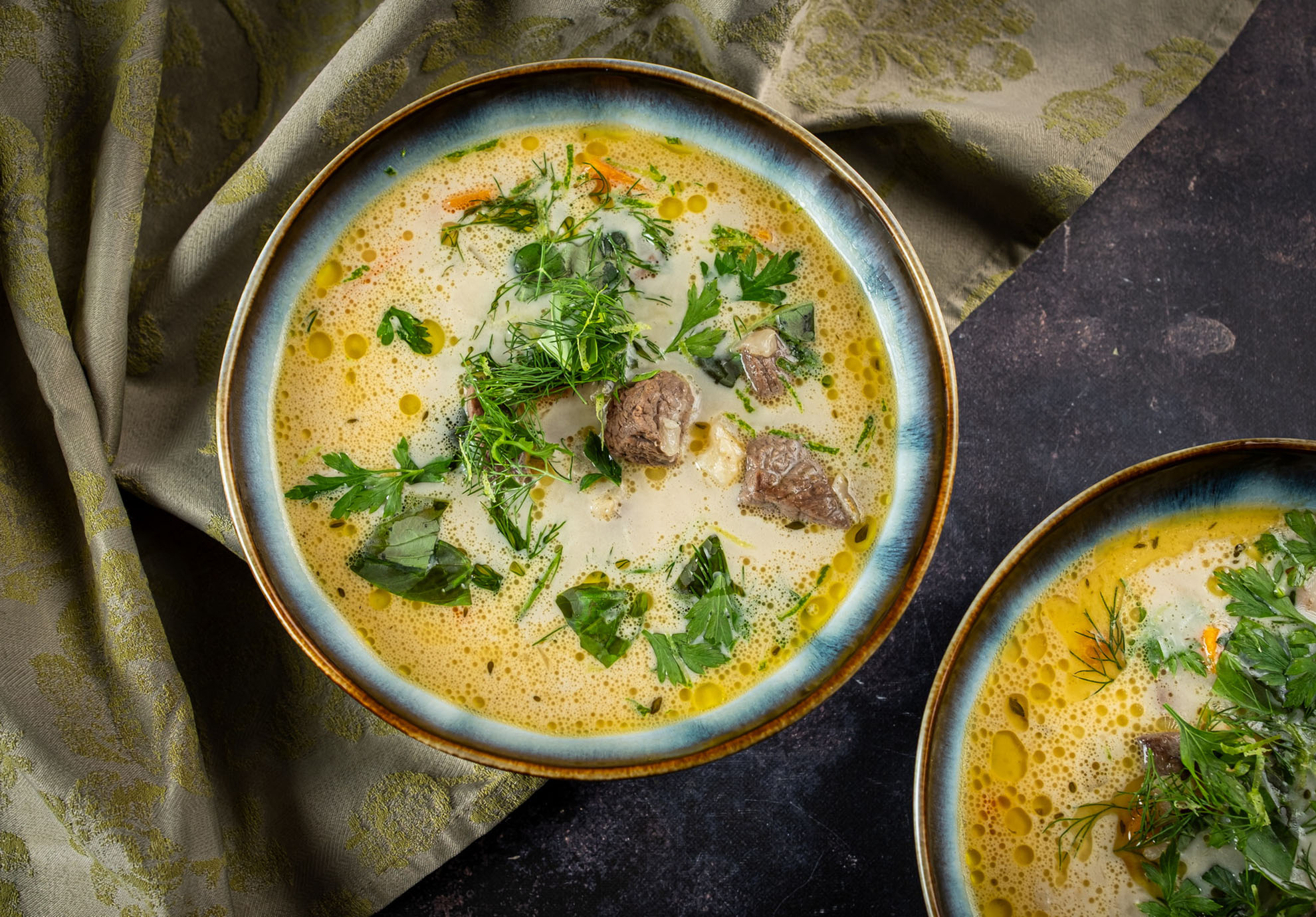 Egy tál krémes leves szarvashús darabokkal, friss fűszernövényekkel, például petrezselyemmel és kaporral díszítve. A leves texturált sötét felületen áll, mintás zöld szövet mellett.